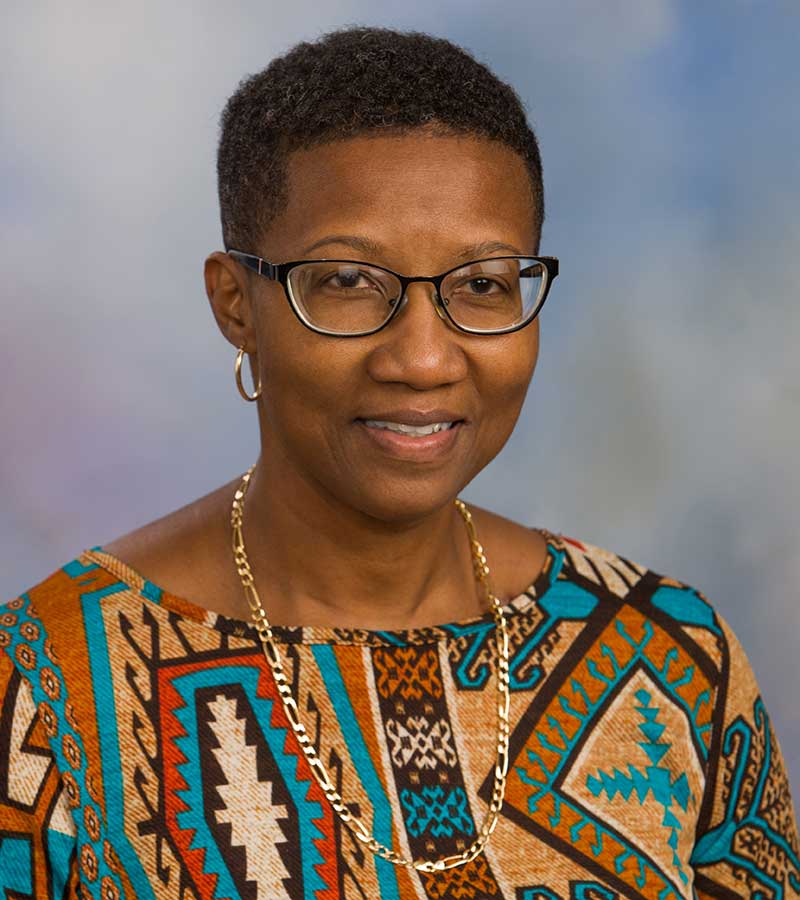 Portrait of Gwen Blakely with short hair and black framed glasses. She is wearing a gold chain necklace and a colorful top. 