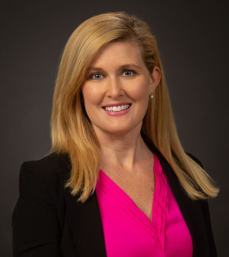 Portrait of Alaina Herrington with long hair wearing a black blazer with a bright pink shirt underneath.
