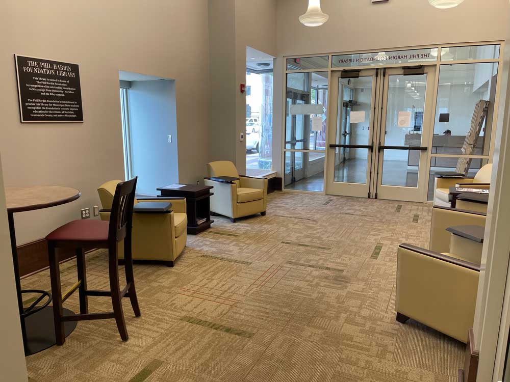 Photo of the sitting area as you enter the Riley Campus Phil Hardin Library. Tan carpet with cream colored chairs with brown swivel desks, a green leafy plant in the corner with a Phil Hardin foundation plaque on the wall with a window looking outside