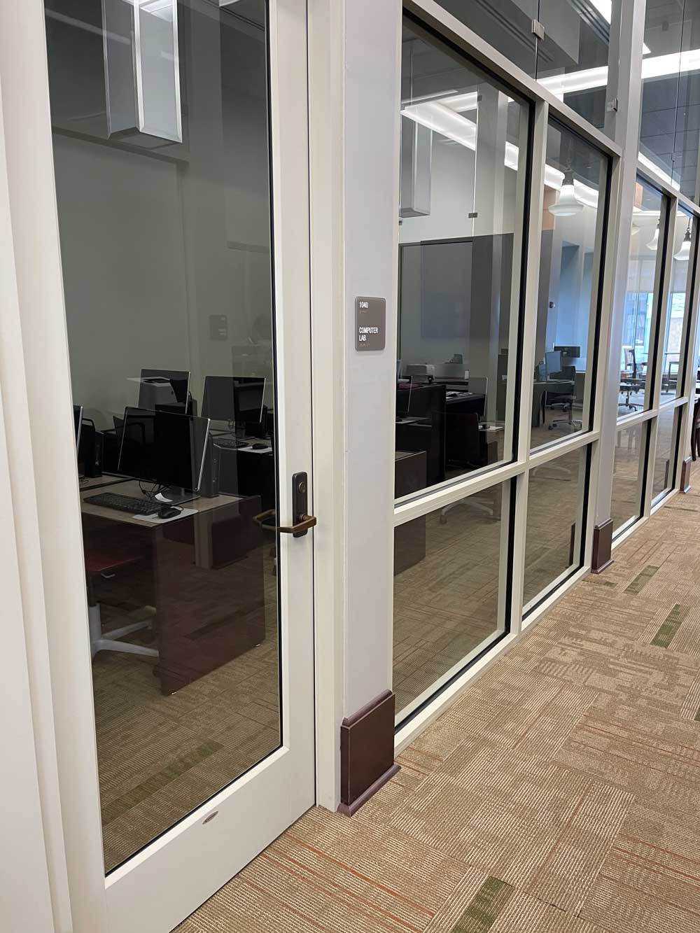 Looking through the glass door and wall into the computer lab where there are rows of tables with black desktop computers and a large tv monitor at the front of the space