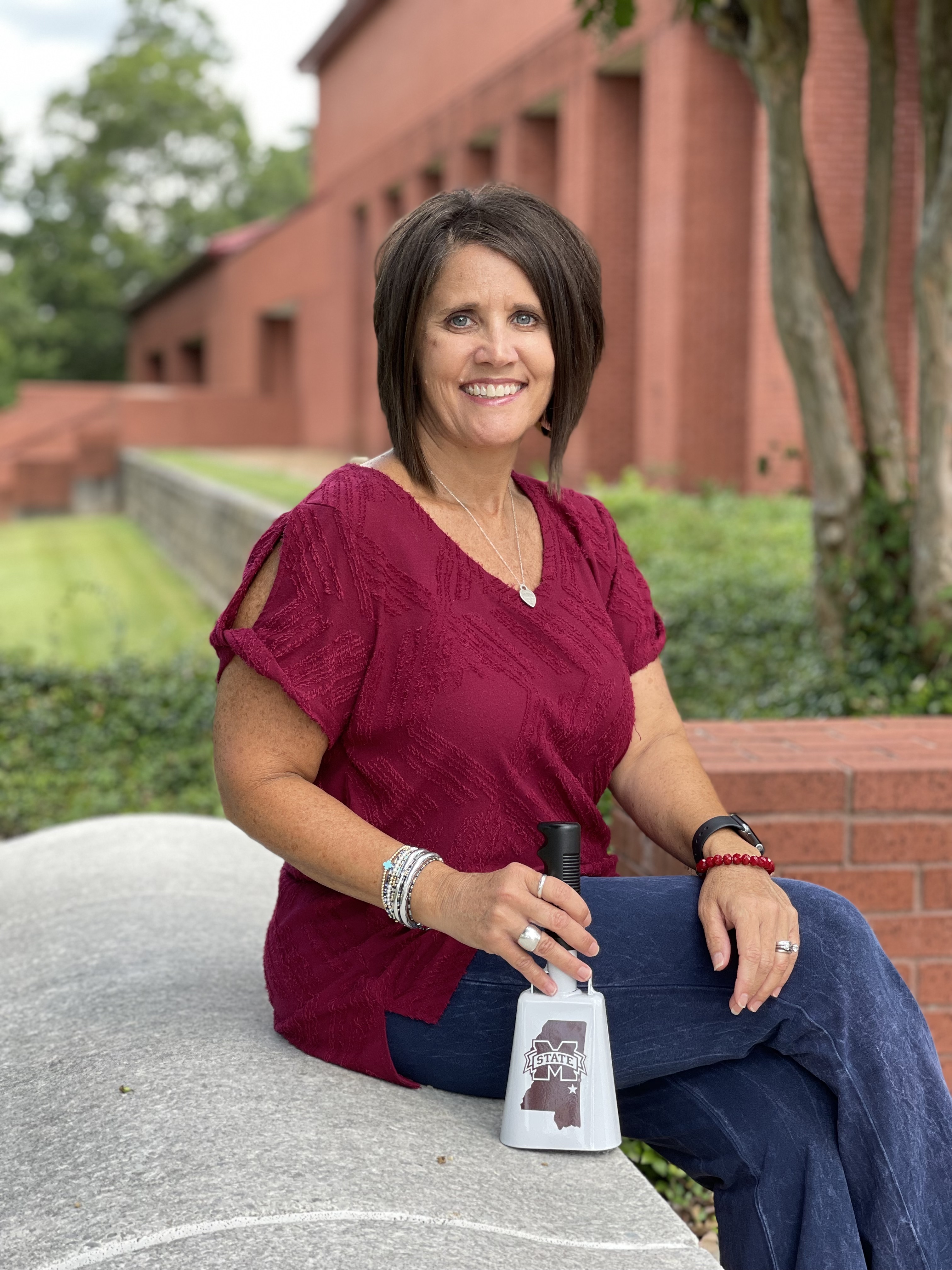 Candy Adams sitting on brick wall wearing maroon shirt and blue pants with cowbell propped in front 