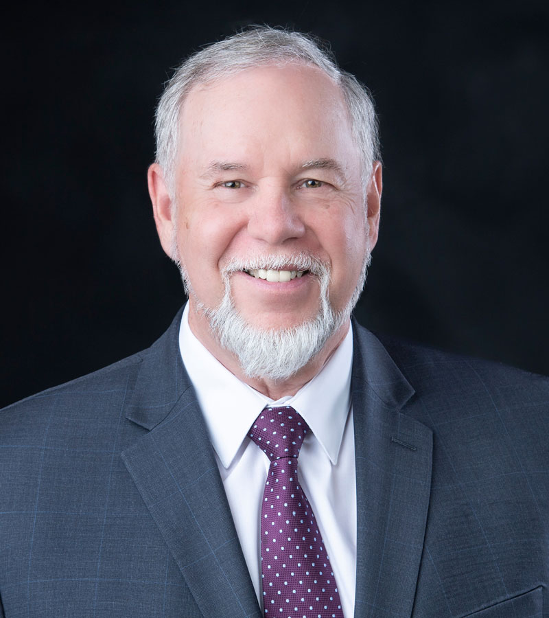 Portrait of Dr. Harold Jones with grey hair and beard wearing a blue blazer, white shirt, and maroon tie. 
