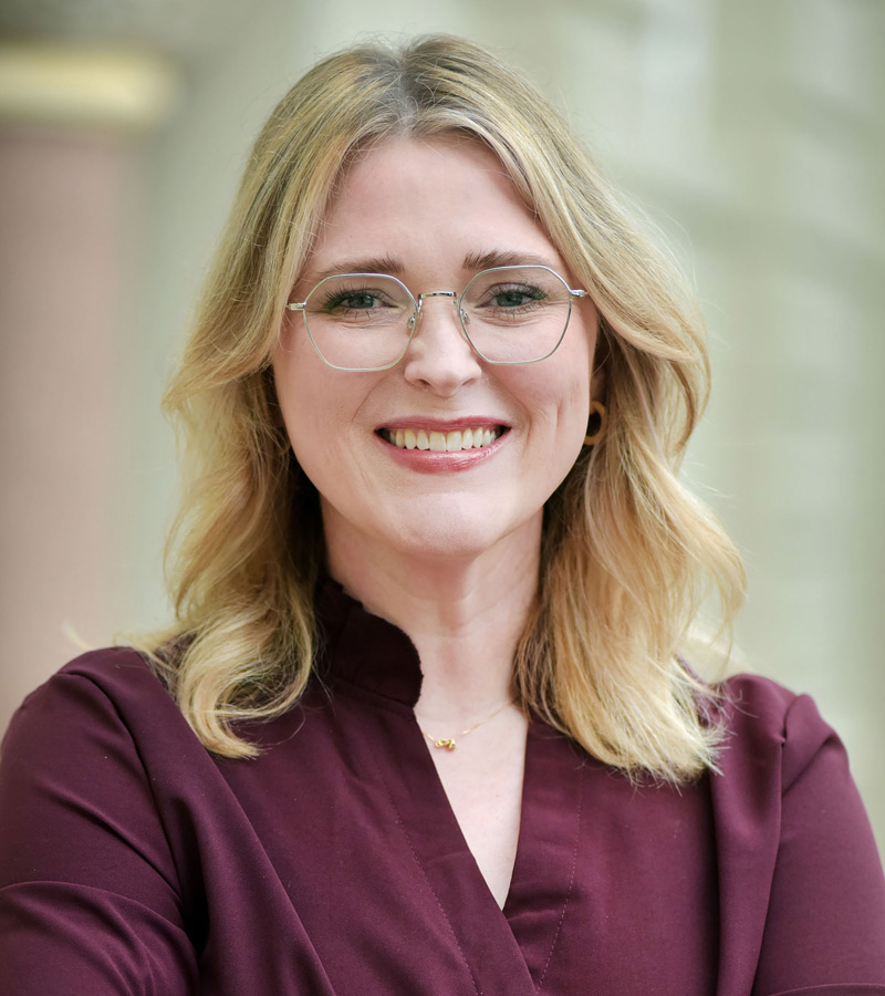 Portrait of Kayla Carr with blonde hair and glasses wearing a maroon top. 