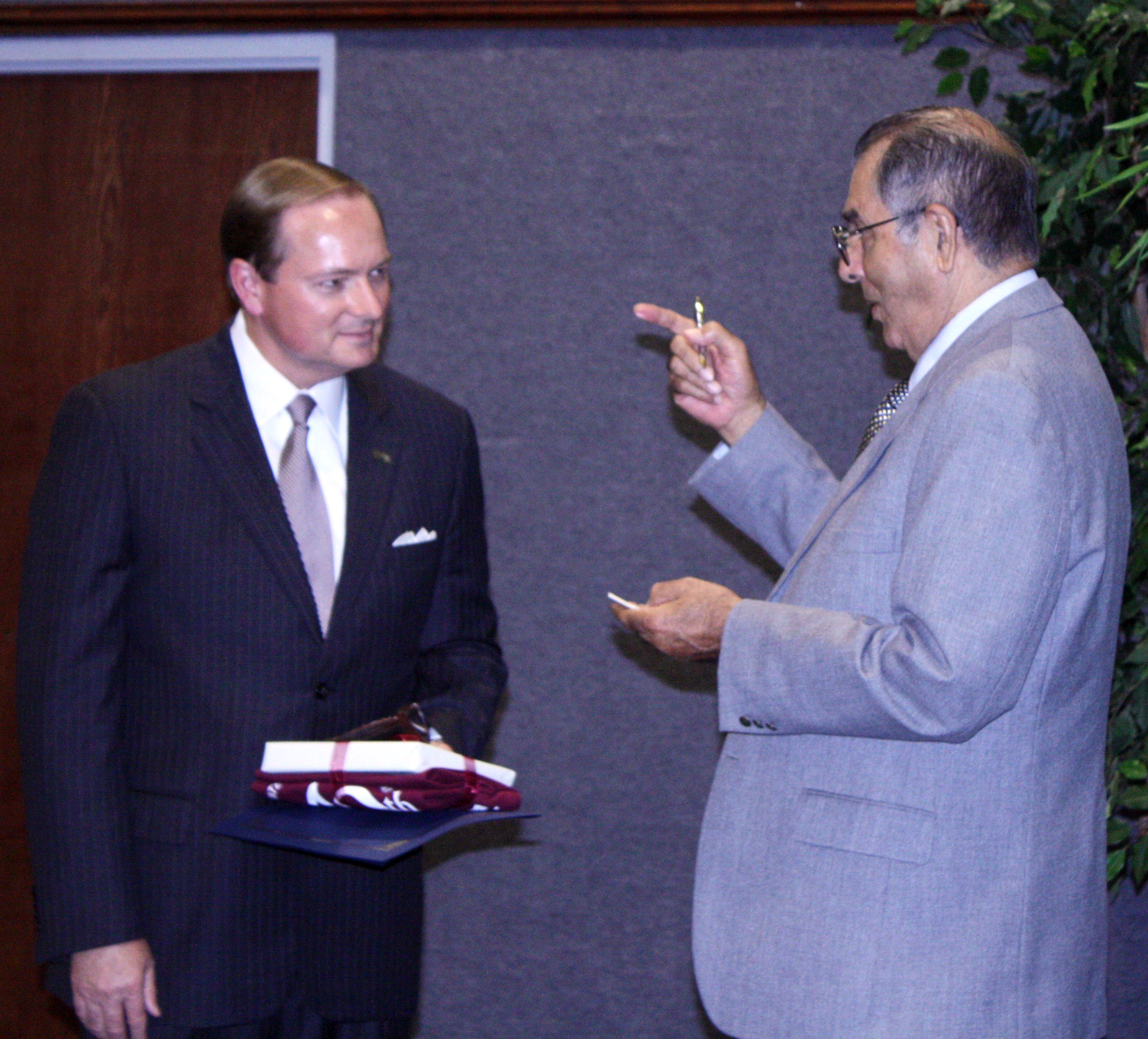 Balding middle age man wearing gray suit and maroon tie speaks with middle eastern older man with glasses