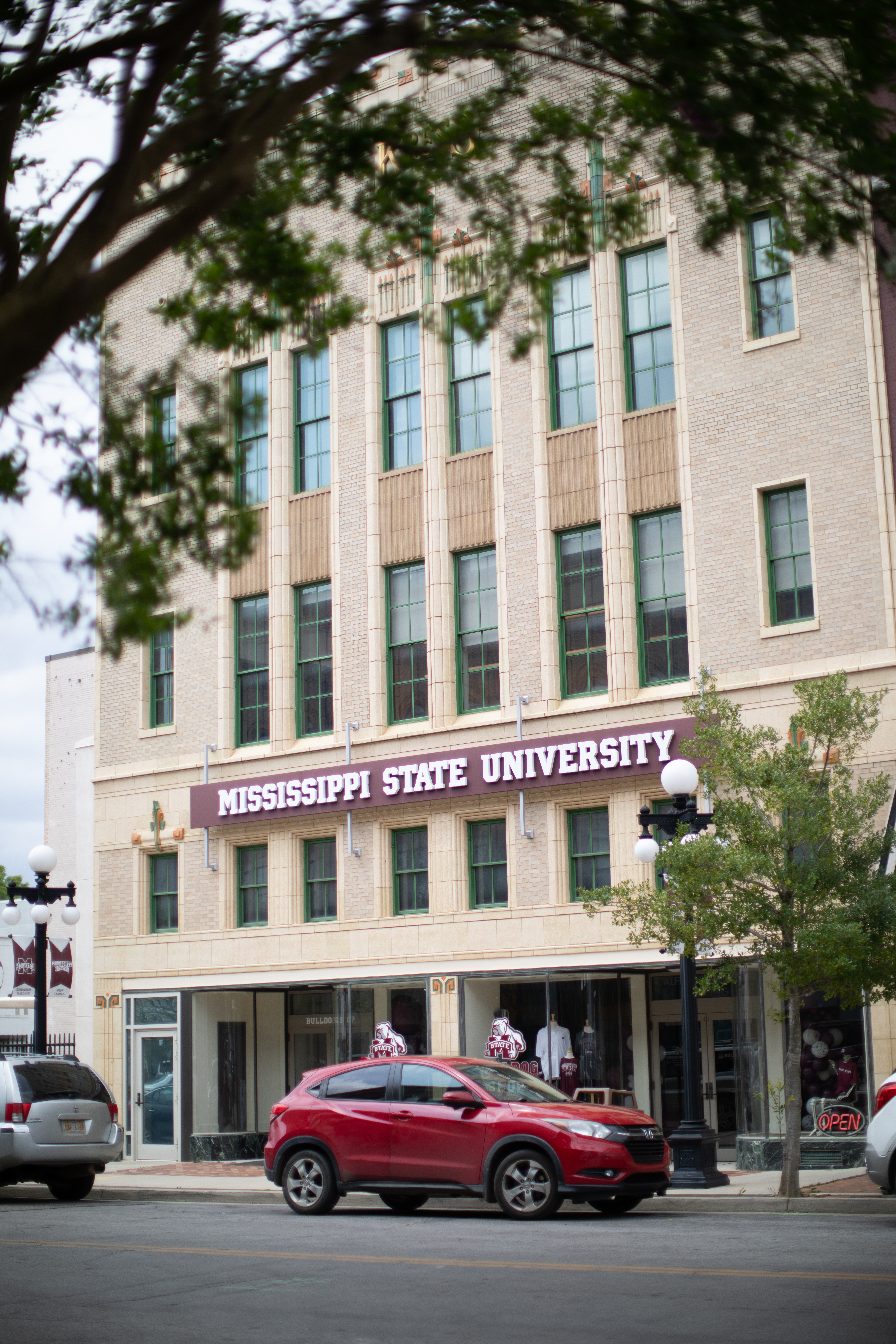 Rosenbaum Building through trees with light brick and tall windows and front sign "Mississippi State University"