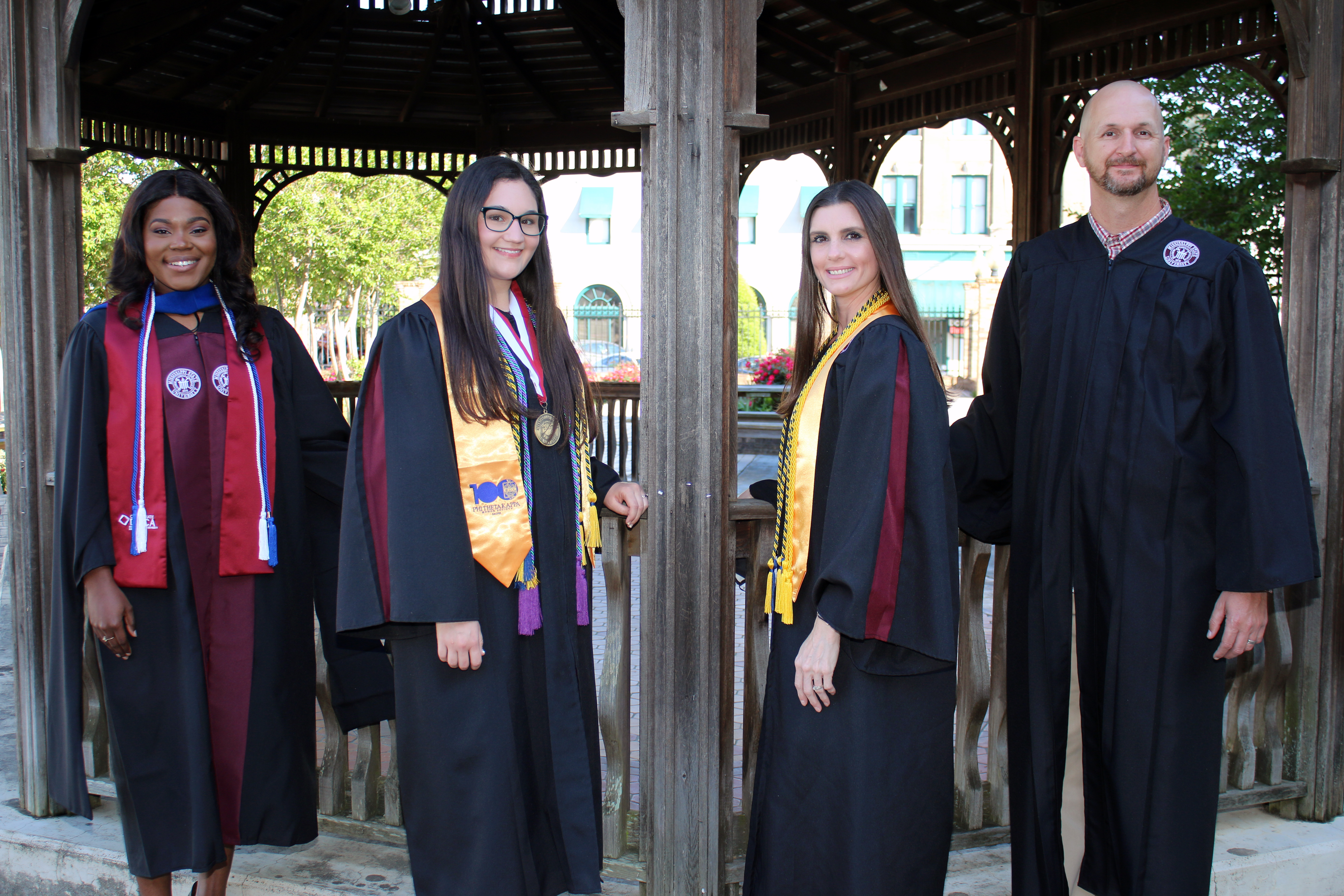 Four students one black female, one white female with glasses, one white female with long black hair and one white tall male all in graduation regalia