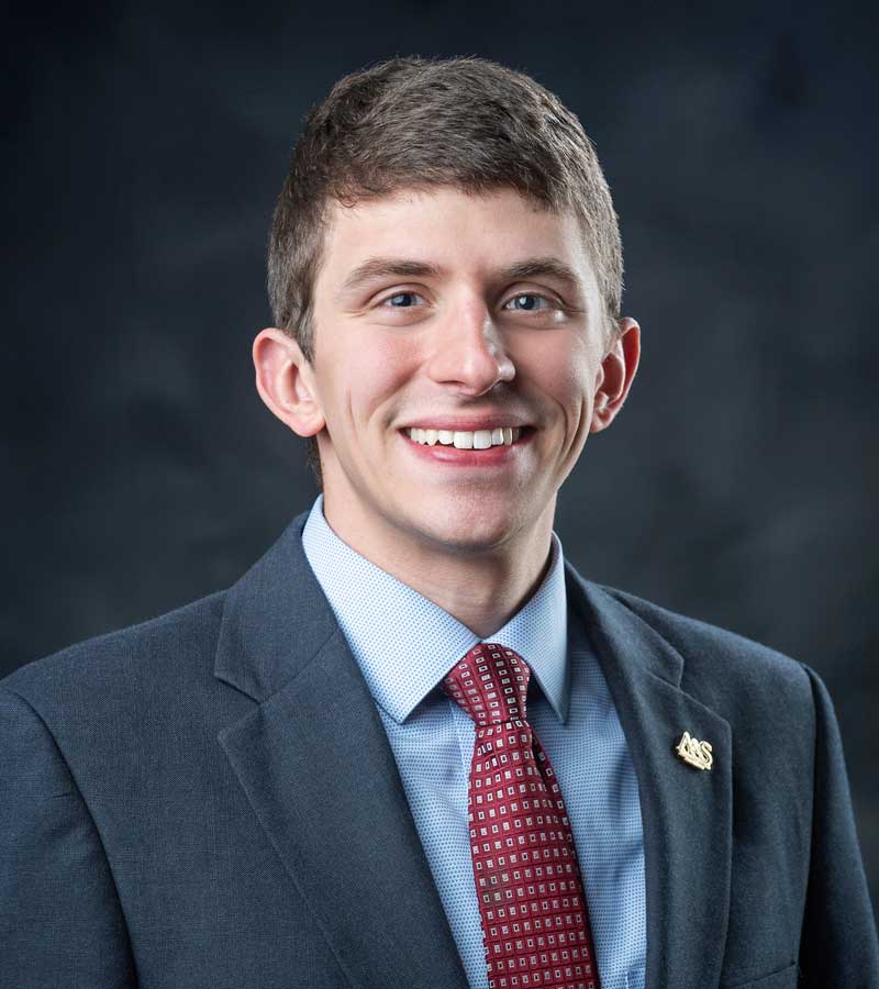 Portrait of Ben Pace wearing a blue blazer with a light blue collared button down shirt and a maroon tie with a gold lapel pin.