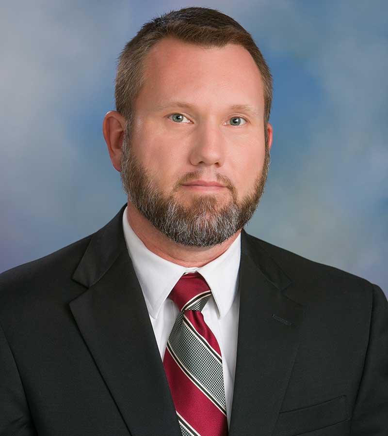 Portrait of Andy Rainey with short hair and a beard, wearing a black suit jackwt with a white shirt and a striped tie. 