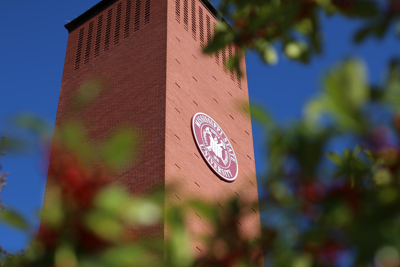 College Park campus brick tower with MSU seal seen through green leafy trees.