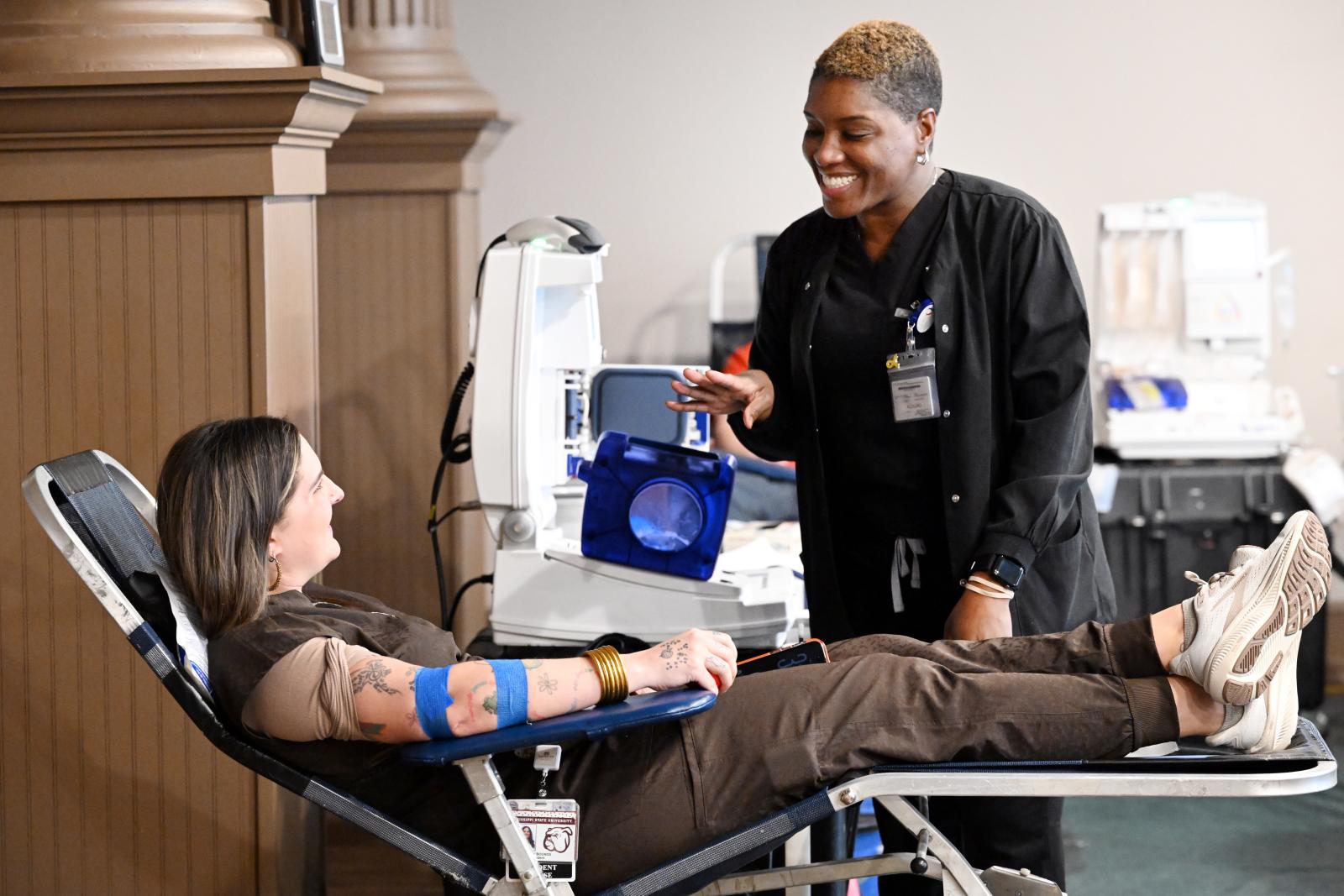 Photo of person in chair (blood donor) talking to another person who is standing next to chair.