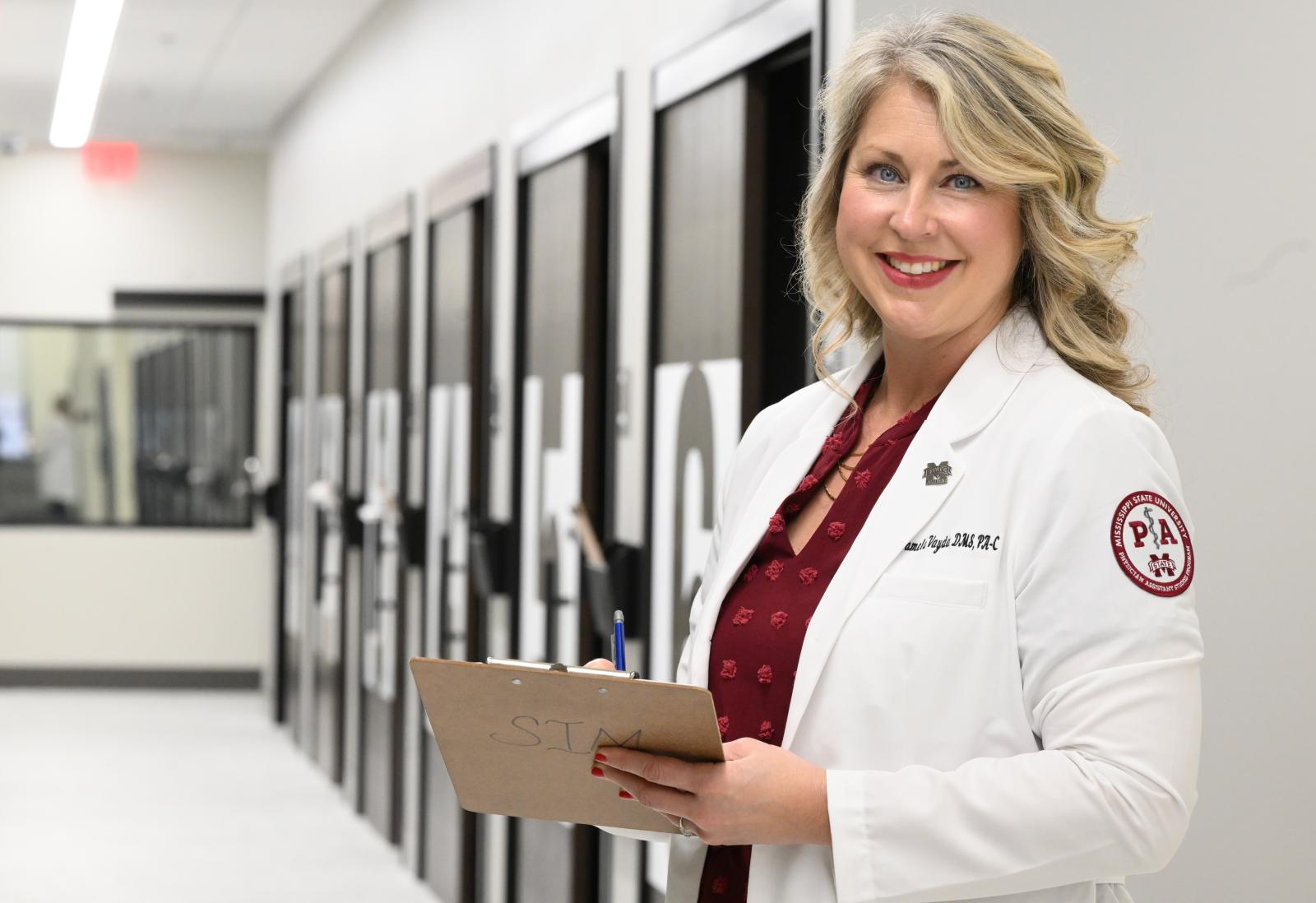 Photo of Pam Vayda in white coat holding a clipboard and smiling at camera in front of long hallway