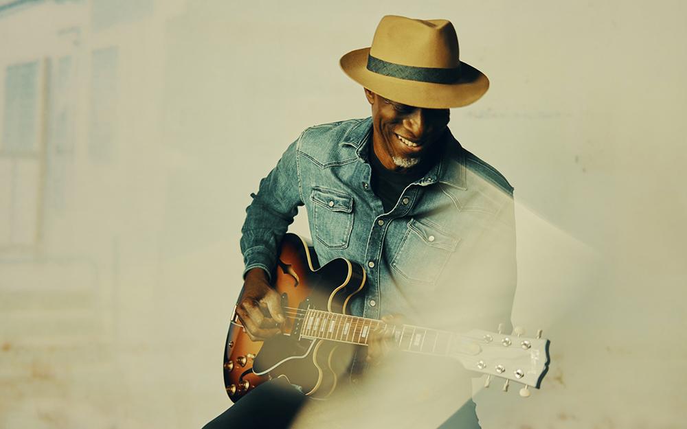 Picture of Keb Mo wearing a tan hat a denim shirt and holding a brown guitar.